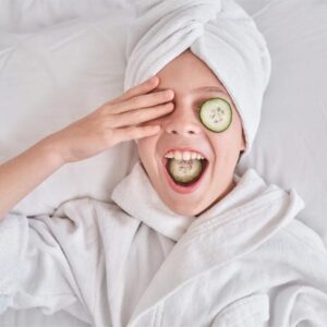 girl smiling with her hair in a towel and a cucumber on her eye to relax